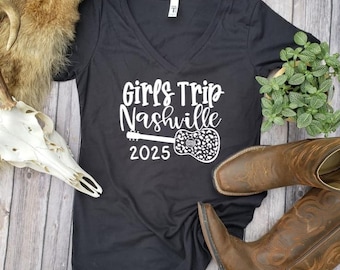 May include: Black v-neck t-shirt with white text that reads "Girls Trip Nashville 2025" and a white silhouette of a guitar with a leopard print design.