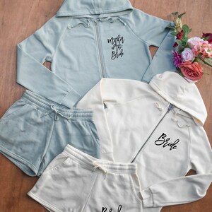 May include: A collection of matching bridal apparel in soft pastel colors. Includes a light blue zip-up hoodie with "Mother of the Bride" text, a white zip-up hoodie and shorts with "Bride" text, and matching shorts. A bouquet of flowers is in the background.