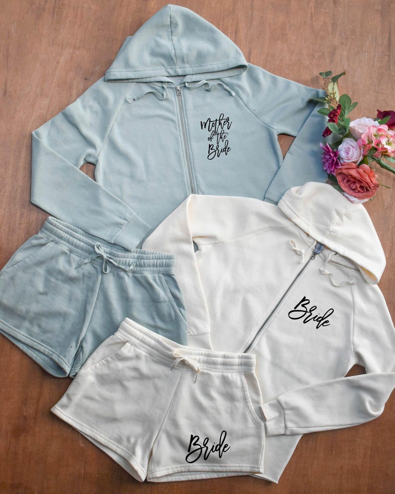 May include: A collection of matching bridal apparel in soft pastel colors. Includes a light blue zip-up hoodie with "Mother of the Bride" text, a white zip-up hoodie and shorts with "Bride" text, and matching shorts. A bouquet of flowers is in the background.