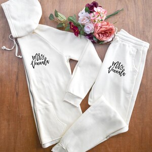 May include: Cream-colored matching sweatsuit set with the text "Mrs. Vennala" in black script. The set includes a zip-up hoodie and sweatpants. A bouquet of pink, red, and white flowers rests near the clothing.