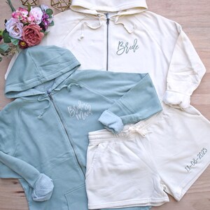 May include: An assortment of matching bridal party apparel. Includes a cream-colored zip-up hoodie with "Bride" script, a blue-green zip-up hoodie with "Bridesmaid" script, and cream-colored shorts with the date "11-08-2025".