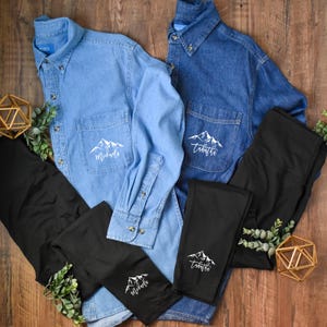 May include: Two long-sleeve denim button-down shirts in light and dark blue, paired with two pairs of black leggings. Each shirt and legging features a white graphic of a mountain range with the names Michaela or Tabatha printed below.