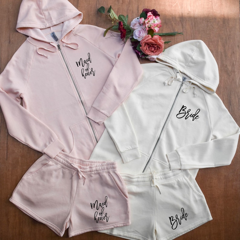 May include: A light pink zip-up hoodie and matching shorts set with "Maid of Honor" printed on the front. An off-white zip-up hoodie and matching shorts set with "Bride" printed on the front. A bouquet of flowers is in the background.