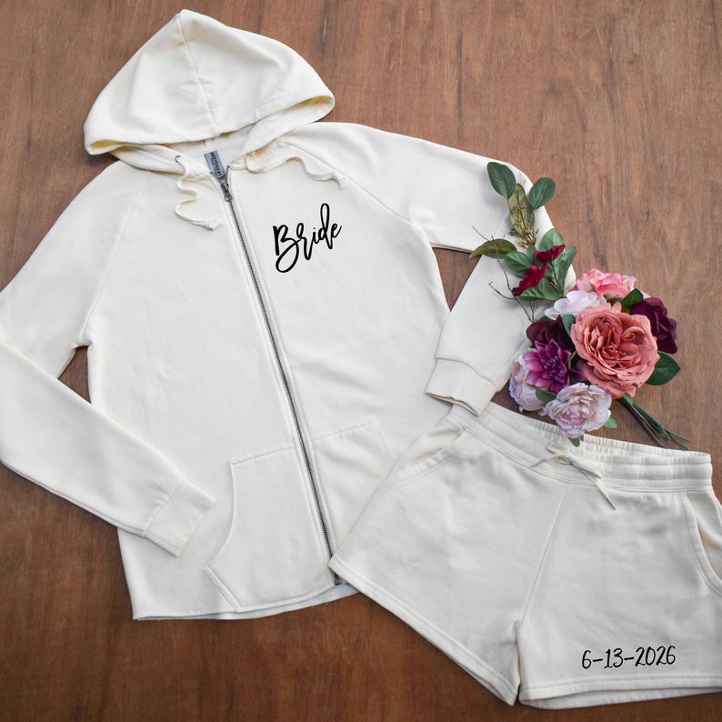 May include: Cream-colored zip-up hoodie and matching shorts set. The hoodie features the word "Bride" in black script and a front zipper. The shorts have a drawstring and the date "6-13-2026" printed on them. A bouquet of flowers is next to the set.
