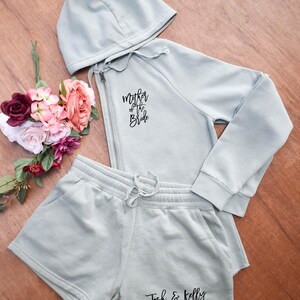 May include: A light green hoodie and shorts set. The hoodie has a zipper and the text "Mother of the Bride". The shorts have the text "Josh & Kelly". A bouquet of flowers is next to the set.