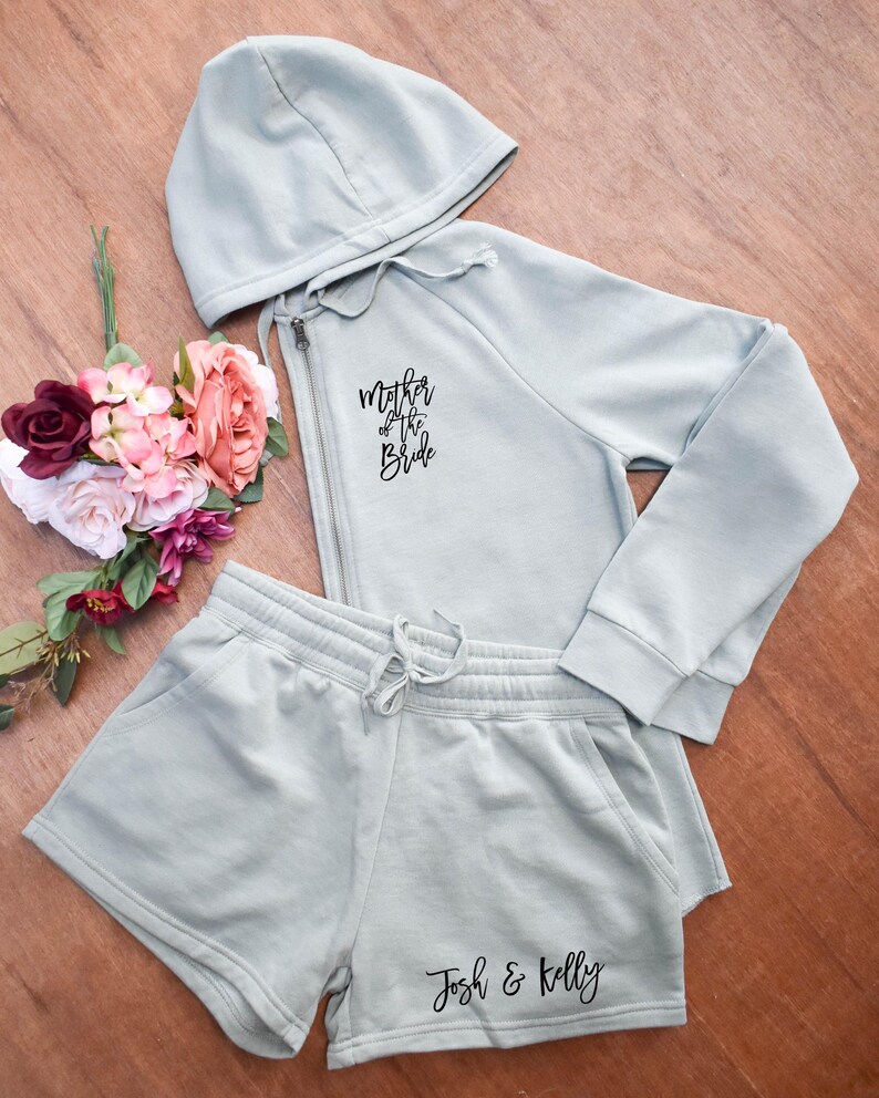 May include: A light green hoodie and shorts set. The hoodie has a zipper and the text "Mother of the Bride". The shorts have the text "Josh & Kelly". A bouquet of flowers is next to the set.
