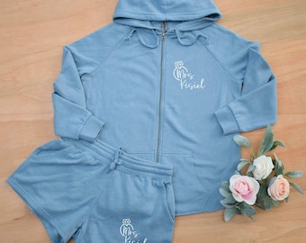 May include: A light blue zip-up hoodie and matching shorts set. The hoodie has a hood and the text "Mrs. Kisiel" with a diamond ring graphic. The shorts also feature the same text and graphic. A bouquet of white and pink roses is in the background.