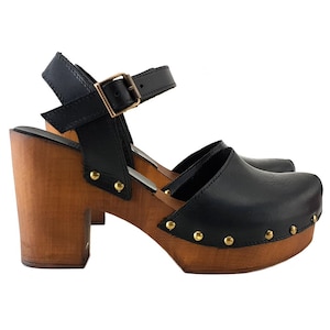 Puede incluir: Zuecos de tacón de cuero negro con plataforma de madera y correa de tobillo. Los zuecos tienen tachuelas doradas alrededor del borde de la plataforma.