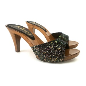 Puede incluir: Zuecos de tacón alto con base y tacón de madera. El diseño de punta abierta presenta una correa de purpurina negra. Los tacones miden aproximadamente 10 cm de alto. La marca "Kiara Shoes" es visible en la plantilla.