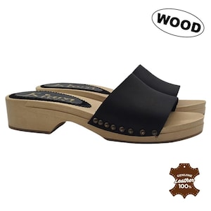 Könnte beinhalten: Schwarze Lederclogs mit Holzsohlen. Die Clogs haben ein Slip-on-Design mit einem breiten Riemen. Die hellbraunen Sohlen kontrastieren mit dem schwarzen Obermaterial. Das Wort "WOOD" steht in einem weißen Oval. Die Clogs sind mit "Genuine Leather 100%" gekennzeichnet.