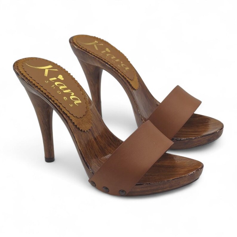 Puede incluir: Un par de zuecos de tac&oacute;n de cuero marr&oacute;n con plataforma de madera y tac&oacute;n alto. Los zuecos tienen una sola correa en la parte superior del pie y est&aacute;n decorados con peque&ntilde;os tachuelas met&aacute;licas. El nombre de la marca "Kiara Shoes" est&aacute; impreso en la plantilla.
