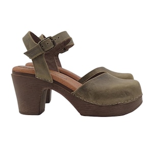 Könnte beinhalten: Ein Paar olivgrüne Ledersandalen mit Absatz, Knöchelriemen und Schnalle. Die Sandalen haben eine Holzplattform und einen dicken Absatz.