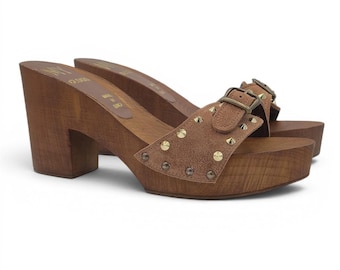 May include: A pair of brown suede mules featuring a wooden platform sole and block heel. The upper strap includes a decorative buckle and gold-tone metal studs. The interior footbed is stamped with the text Made in Italy and a brand logo.