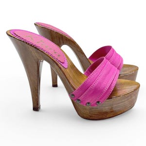 Puede incluir: Un par de zuecos de tacón alto con plataformas y tacones de madera. Los zapatos presentan una correa y una plantilla de color rosa brillante, con una base de madera marrón oscuro. La plantilla tiene texto en un diseño rosa y morado.