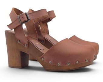 May include: A pair of brown leather clogs with wooden heels. The clogs have an ankle strap with a buckle and are adorned with metal studs along the sole. The heels are approximately 7.6 cm high.