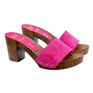 May include: A pair of pink suede clogs with wooden heels and a gold metal accent. The clogs are made in Italy and have the brand name "MY BLOGS" on the side.