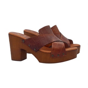 May include: Brown leather platform clogs with a criss-cross strap design and studded accents. The clogs have a wooden platform and heel.