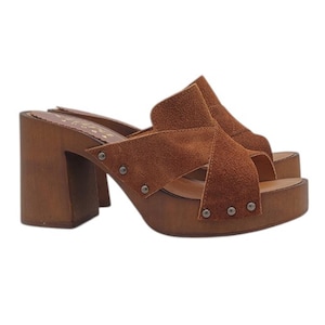 May include: Brown suede heeled clogs with a wooden platform and a criss-cross strap design. The clogs have metal studs on the straps and platform.