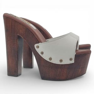 Könnte beinhalten: Holz-Plateau-Clogs mit hohem Absatz und weißem Lederriemen. Die Clogs haben eine dunkelbraune Holzbasis und einen Absatz, und der Riemen ist mit kleinen, runden Nieten befestigt. Das Plateau misst etwa 5 cm.