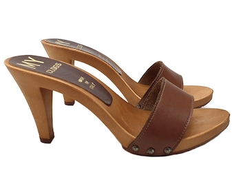 May include: Brown leather heeled clogs with a wooden platform and a single strap across the top. The clogs have a gold metal accent on the strap and are made in Italy.