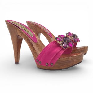 May include: A pair of high-heeled clogs with a wooden base and heel. The clogs feature a bright pink strap adorned with purple and clear gemstones. The footbed is lined with pink fabric and gold trim. The heels are approximately 4 inches tall.