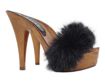 May include: A pair of wooden platform clogs with a high heel and a black fur pom-pom on the top of the shoe.