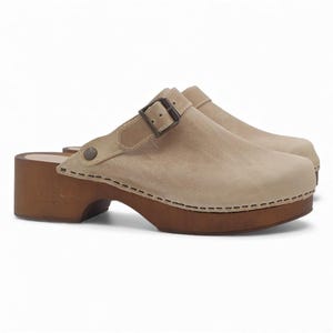 May include: A pair of beige clogs with a wooden sole and heel. The clogs have a leather upper with a buckle closure and decorative stitching. The heel is approximately 3 inches high.