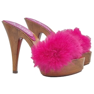 May include: A pair of high-heeled clogs with a wooden base and heel. The clogs feature a vibrant fuchsia faux fur strap across the top. The insole has a pink scalloped edge and a printed design. The heels are approximately 4 inches tall.