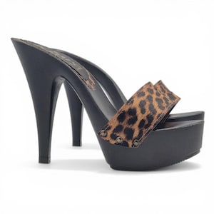 May include: Black platform high heel sandals with a leopard print strap. The sandals have a wooden sole and a stiletto heel. The strap is secured with small silver-colored rivets. The shoes are set against a white background.