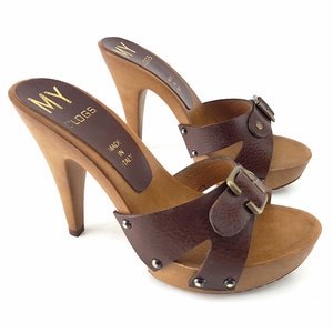 May include: Brown leather heeled clogs with a buckle strap and wooden platform. The clogs are made in Italy and have the brand name "MY CLOGS" printed on the insole.
