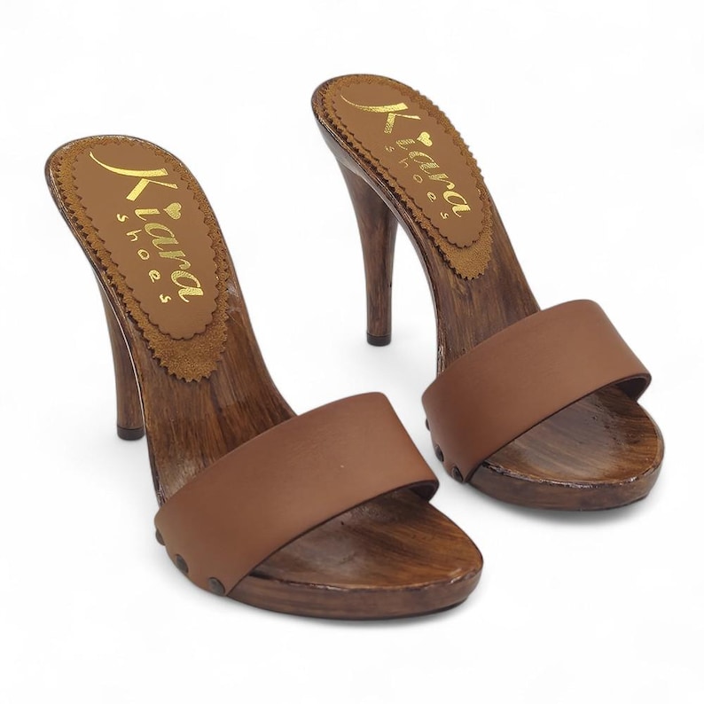 Puede incluir: Un par de zuecos de tac&oacute;n de cuero marr&oacute;n con plataforma y tac&oacute;n de madera. Los zuecos tienen una sola correa en la parte superior del pie y el nombre de la marca "Kiara Shoes" est&aacute; impreso en la plantilla.