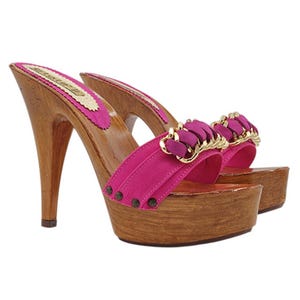 May include: A pair of high-heeled clogs with a wooden base and heel. The shoes feature a bright pink strap with gold-colored accents and a decorative element. The insole is pink with gold trim.