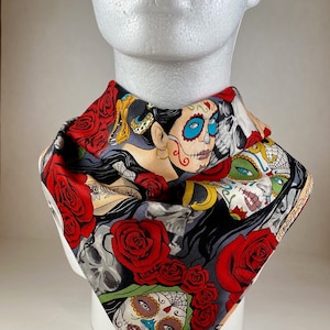 May include: A colorful bandana with a Day of the Dead theme. The bandana features a black and white skull with red roses and a woman with a sugar skull face paint.