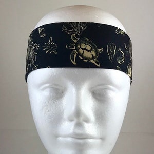 Op de afbeelding: Zwarte hoofdband met een herhaald patroon van goudkleurige zeeschildpadden, schelpen en koraal. De hoofdband is gemaakt van stof en is ontworpen om om het hoofd te worden gedragen. Het ontwerp heeft een nautisch thema.