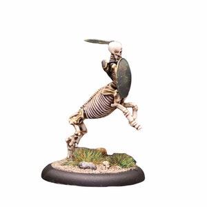 Könnte beinhalten: Eine skelettartige Kentauren-Miniaturfigur mit Schwert und Schild, in Weiß und Braun bemalt, steht auf einem schwarzen Sockel mit grünem und braunem Gras.