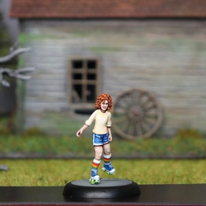 Puede incluir: Una figurita miniatura pintada de una persona joven con cabello rojo que lleva una camisa amarilla, pantalones cortos azules y calcetines a rayas arcoíris. La figurita está sobre patines y está posada como si estuviera patinando.