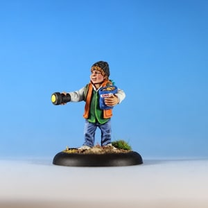 May include: Miniature figure of a person holding a torch and a blue bag labelled "CAFE". The figure wears a black beanie, orange vest, green shirt, and blue jeans. The base is black with a patch of grass.