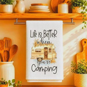 May include: A white waffle-weave kitchen towel featuring a graphic of a vintage camper surrounded by autumn foliage. The text "Life is Better when Camping" is printed in black script and sans-serif lettering above and below the camper.
