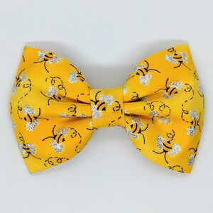 May include: A yellow bow tie featuring a pattern of bees with silver glitter. The bow tie is made from fabric and has a simple design.