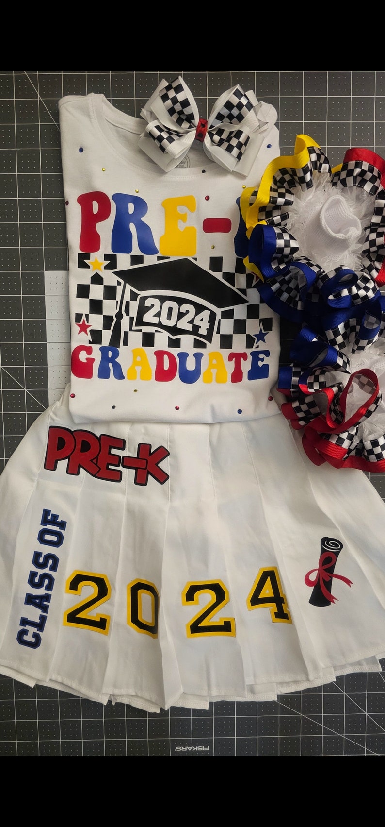 May include: White pre-k graduation outfit with a black and white checkered pattern, red, blue, and yellow accents, and the text "PRE-K 2024 GRADUATE". The outfit includes a white shirt, a white skirt with the text "PRE-K CLASS OF 2024" and a graduation cap.