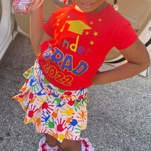 May include: A young girl wearing a red shirt with the text "Kinder Grad 2022" in rainbow letters and a white skirt with colorful handprints. She is wearing rainbow-colored shoes and a sparkly pink purse.