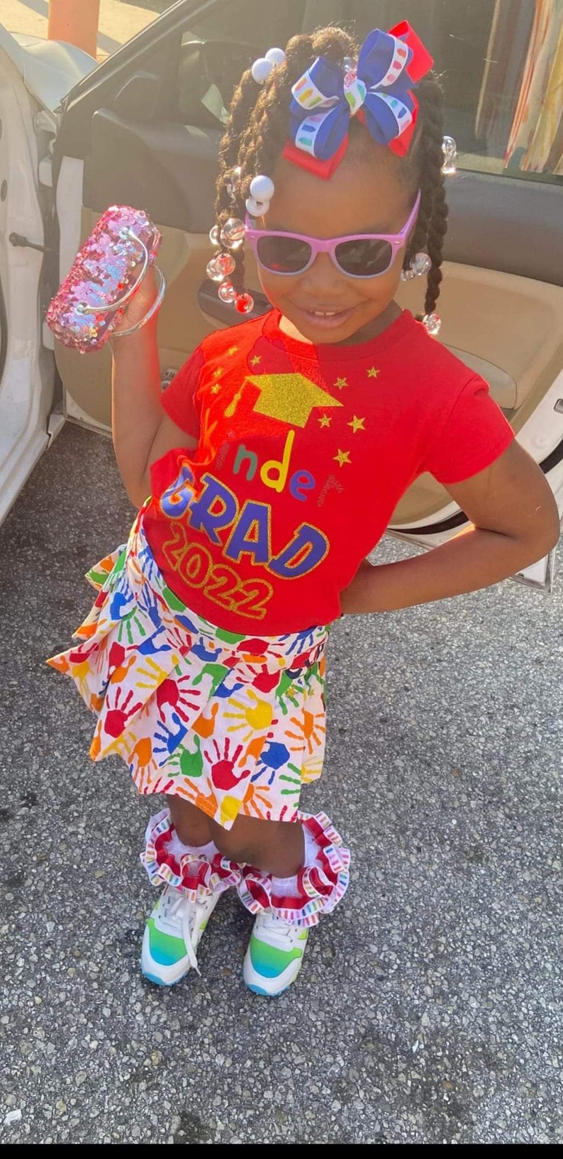 May include: A young girl wearing a red shirt with the text "Kinder Grad 2022" in rainbow letters and a white skirt with colorful handprints. She is wearing rainbow-colored shoes and a sparkly pink purse.