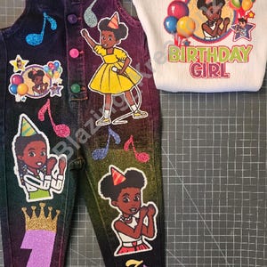 Op de afbeelding: Een donkere spijkerbroek met tuinbroek-bovenstuk in een regenboogverloop, versierd met glitterende muzieknoten, een paars glittercijfer één, een gouden glitterkroon en illustraties van een meisje met een feesthoedje. Inclusief een wit geribbeld shirt met de tekst THE BIRTHDAY GIRL en een bijpassende geel-witte strik.