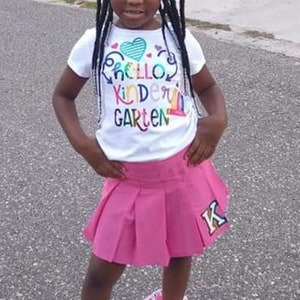 May include: A young girl wearing a white t-shirt with the text "Hello Kindergarten" in rainbow colors and a pink pleated skirt with a gold "K" on the side. She is wearing white sneakers with pink and white ruffles around the ankles and a pink and white bow in her hair.