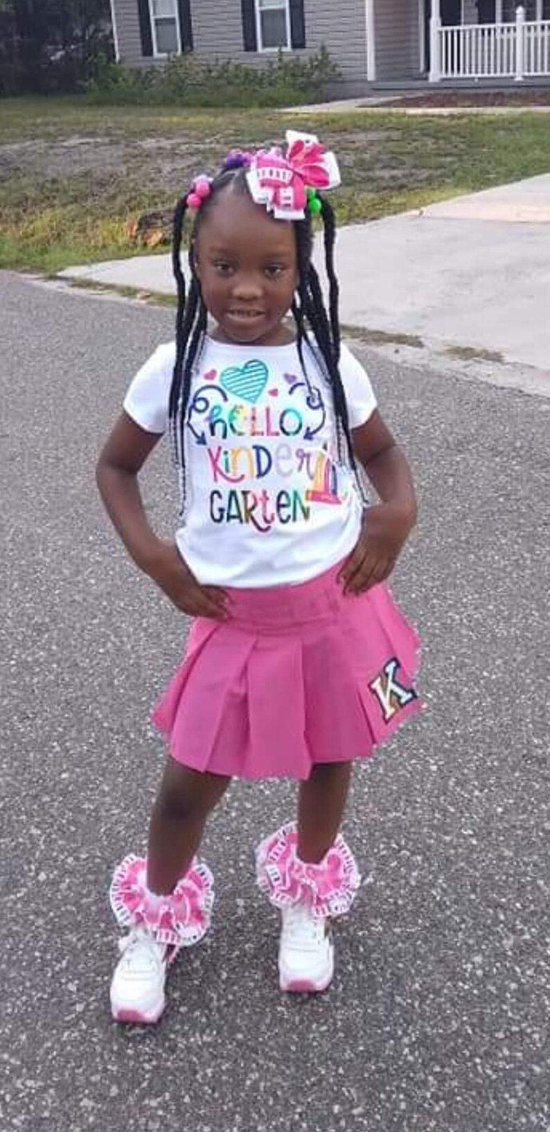 May include: A young girl wearing a white t-shirt with the text "Hello Kindergarten" in rainbow colors and a pink pleated skirt with a gold "K" on the side. She is wearing white sneakers with pink and white ruffles around the ankles and a pink and white bow in her hair.