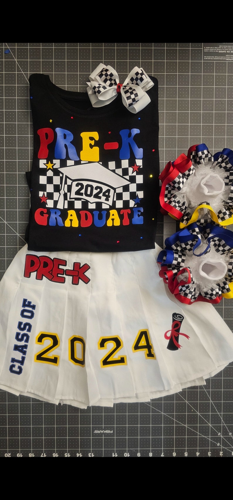 May include: A white pleated skirt with the text "PRE-K CLASS OF 2024" in blue, red, and yellow. The skirt has a red, white, and blue bow with black and white checkered ribbon on the side.