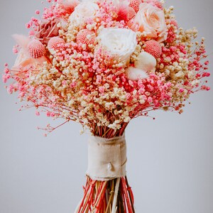 May include: A bouquet of pink and white flowers, including roses and baby's breath, tied with a beige ribbon and a pearl. The stems are wrapped with red, pink, and natural-coloured sticks, set on a wooden base.