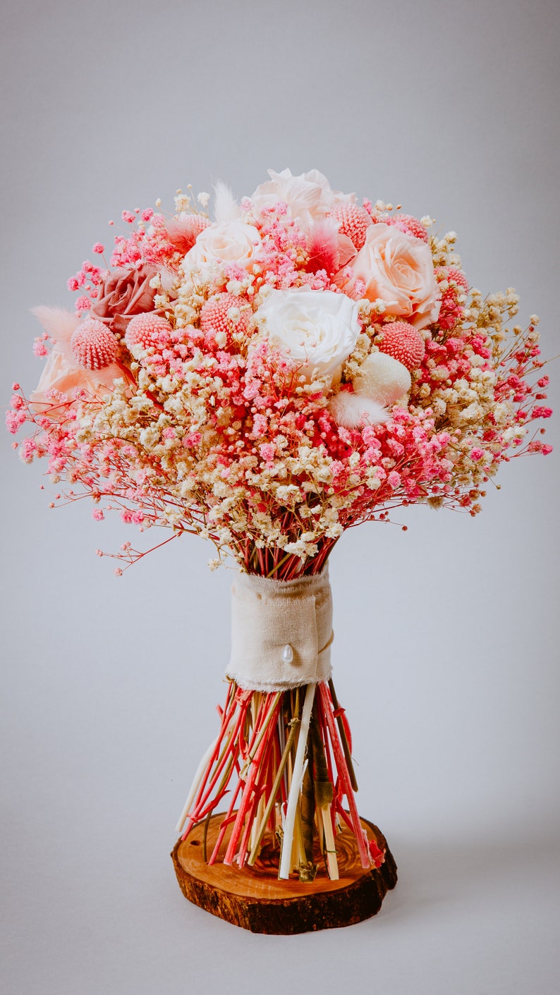 May include: A bouquet of pink and white flowers, including roses and baby's breath, tied with a beige ribbon and a pearl. The stems are wrapped with red, pink, and natural-coloured sticks, set on a wooden base.