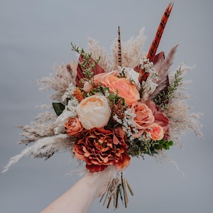 Può includere: Un bouquet di fiori secchi in tonalità pesca, arancio e marrone. Il bouquet comprende erba della pampa secca, rose secche e altri fiori secchi.