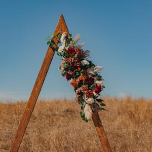 May include: A wooden triangular wedding arch decorated with a floral arrangement. The flowers are a mix of red, orange, and white roses, pampas grass, and greenery. The arch is set against a backdrop of a field of dry grass and a clear blue sky.